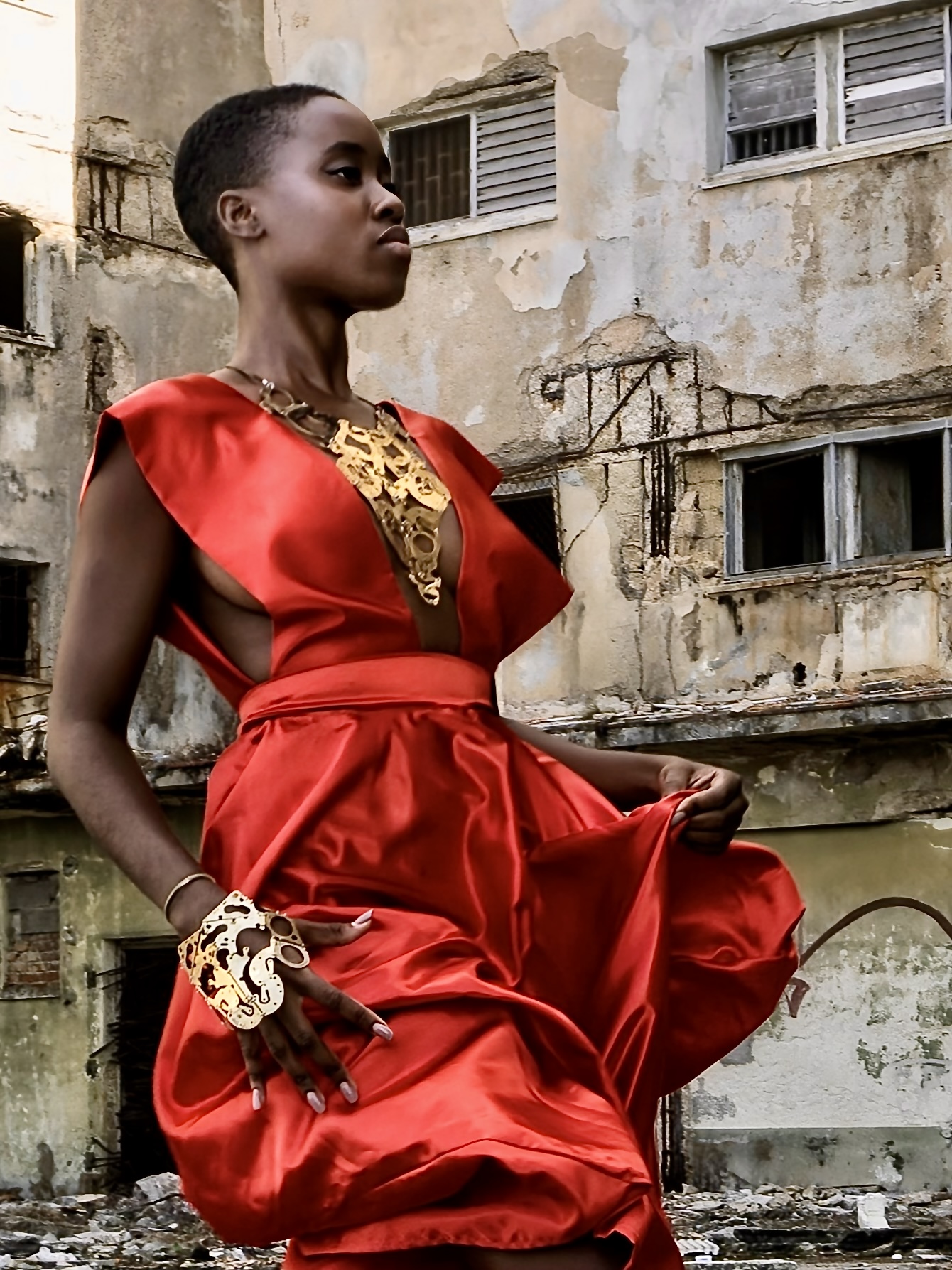 Woman in red dress with gold accessories standing outdoors in front of an old building.
