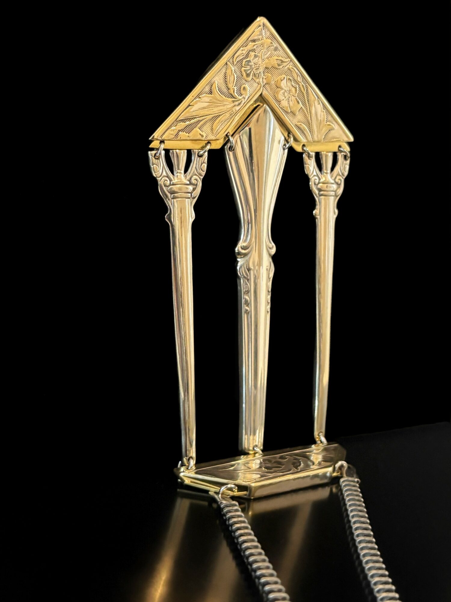 Golden decorative lamp with three tall, ornate columns and a triangular top, on a black background.
