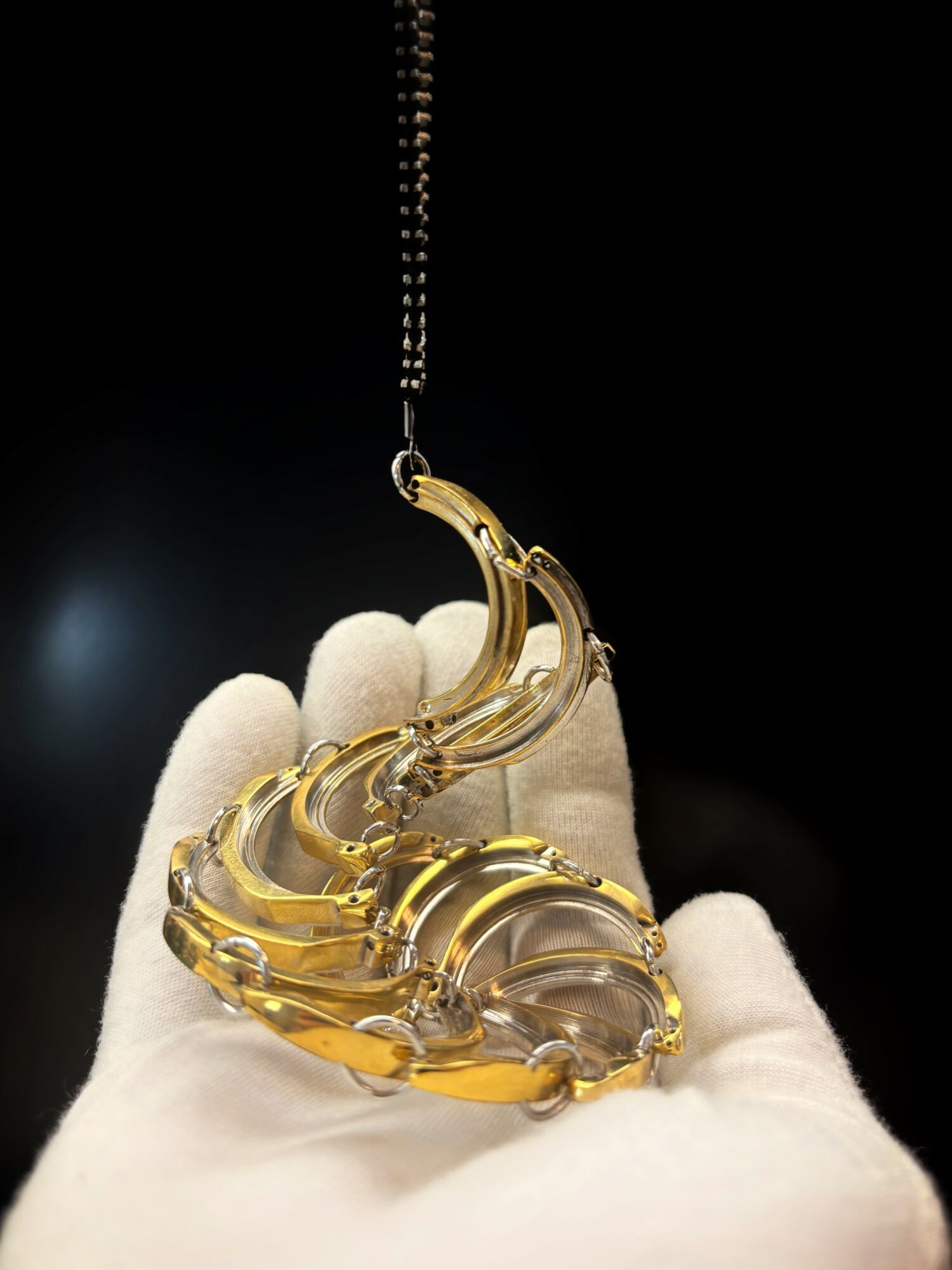 A glass sculpture resembling a crescent moon held by a hand in a white glove, hanging from a chain against a black background.