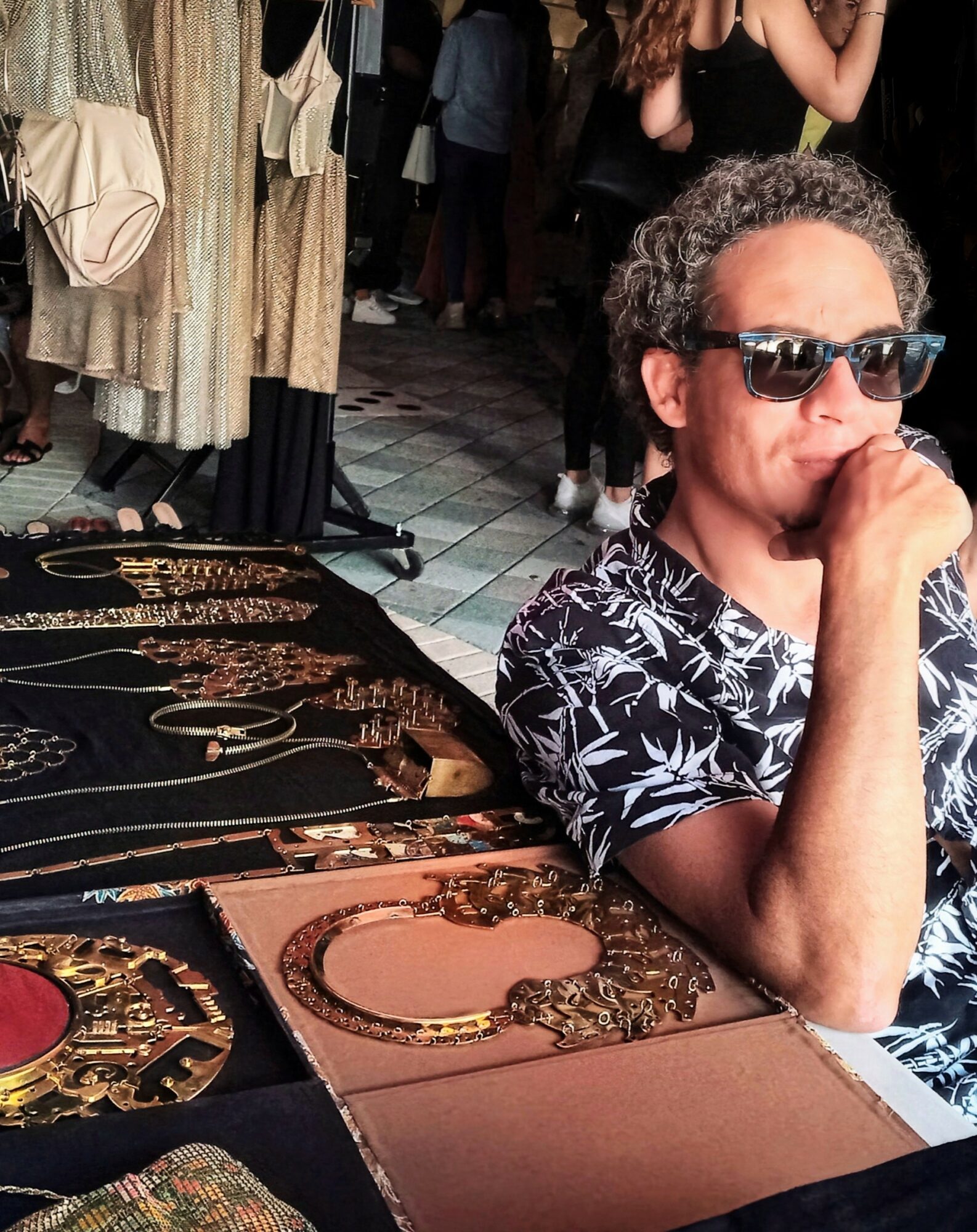 Man with curly hair wearing sunglasses and a patterned shirt sits at a table with jewelry displays, in a busy indoor market.