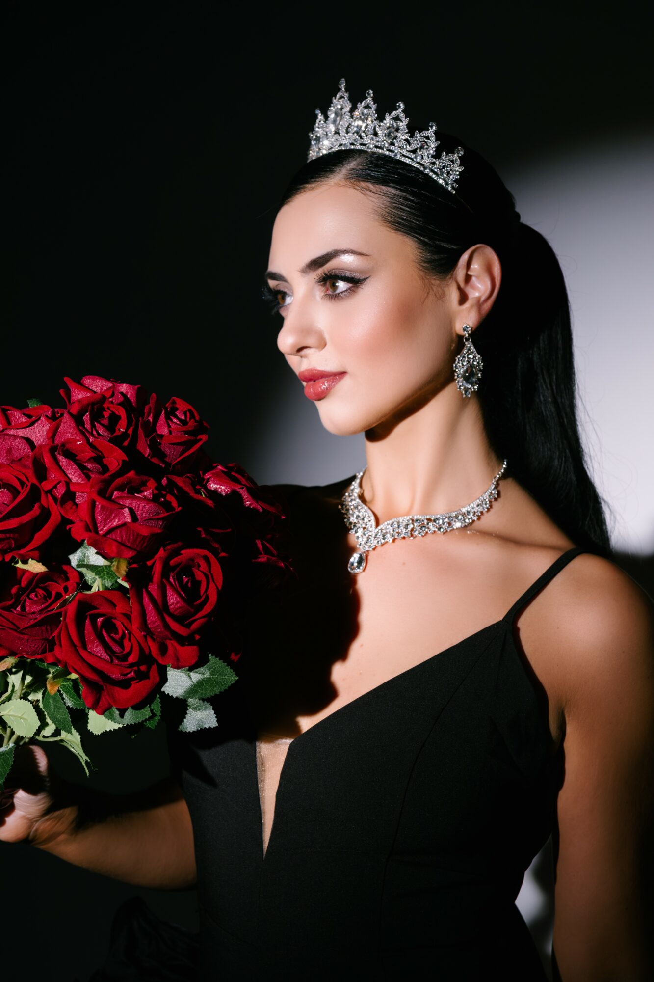 Woman wearing a crown and jewelry holds a bouquet of red roses, dressed in a black gown, against a dark background.