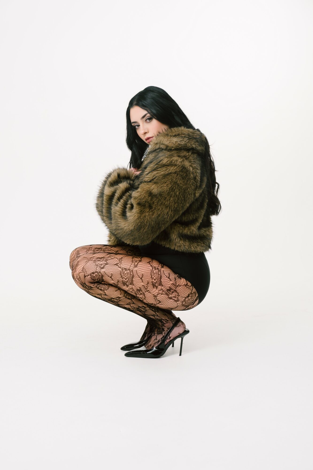 Woman crouching in a fur coat, patterned tights, and high heels against a white background.