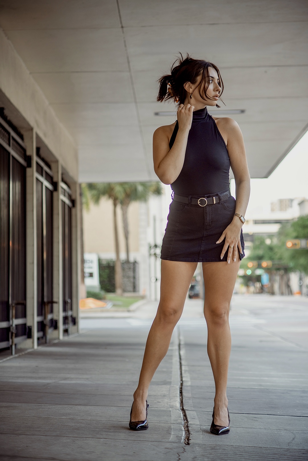 Woman in a black sleeveless dress standing outdoors, looking to the side, with a city street background.