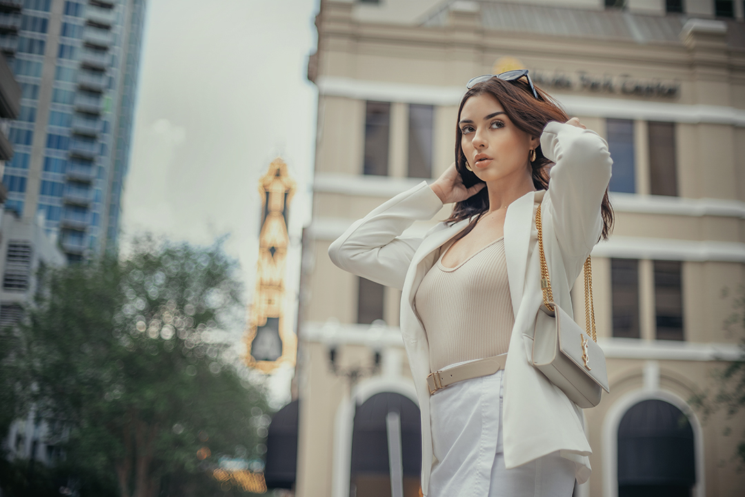 Woman with dark hair in a city street, wearing a white outfit and carrying a beige bag, with buildings in background.