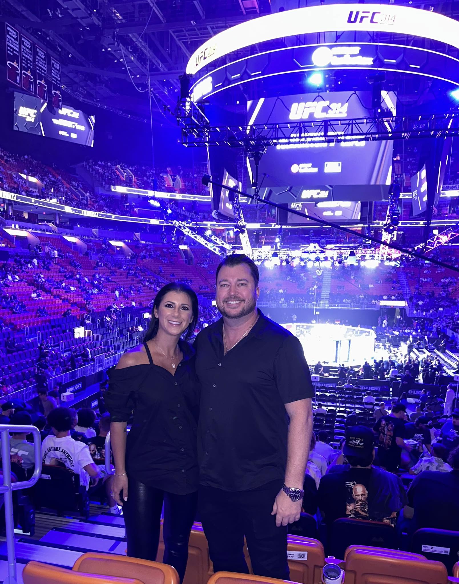 Two people stand in front of a large, brightly lit arena with a crowd and screens displaying UFC branding.