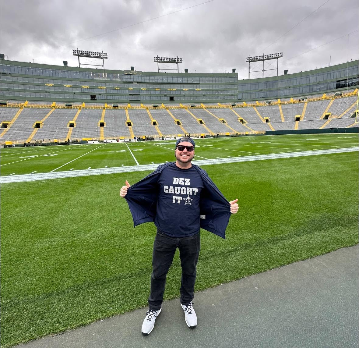 Person standing on a football field in a stadium, smiling, wearing sunglasses, a dark sweatshirt, and white shoes.