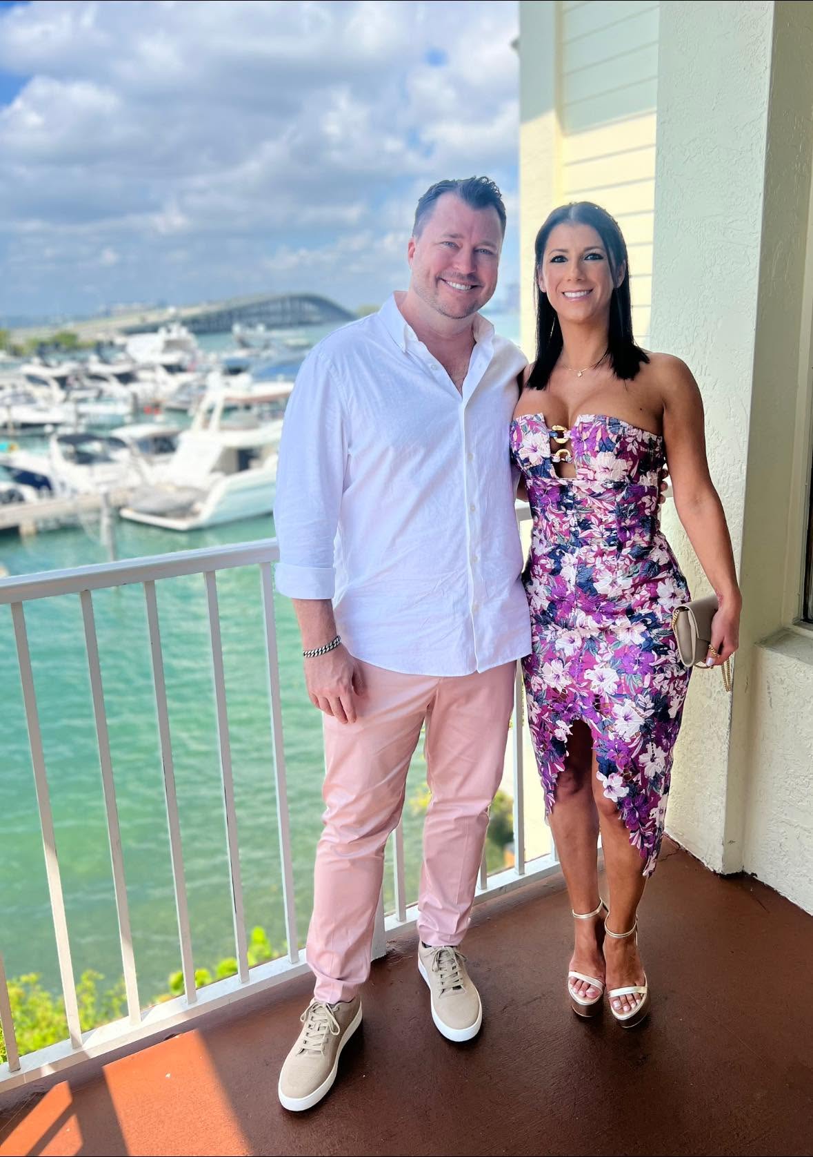 A man and woman stand on a balcony with a marina and boats in the background, smiling at the camera.