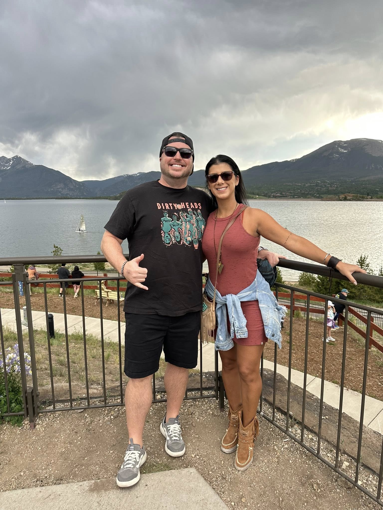 A man and woman stand together outdoors near a lake with mountains in the background, smiling and posing for the photo.