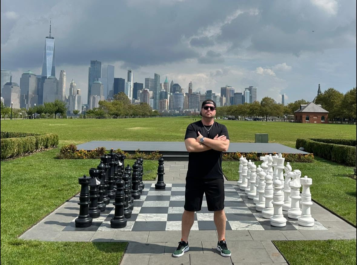 Person standing with arms crossed on a large outdoor chessboard, city skyline in background, cloudy sky, grassy park area.