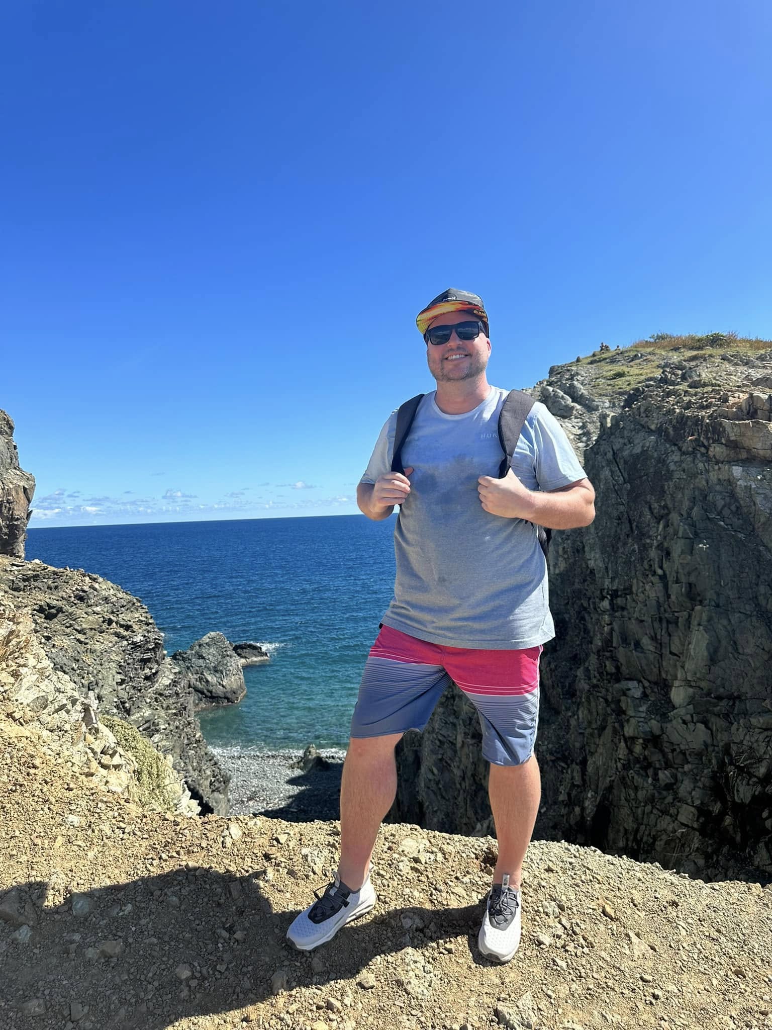 Person standing outdoors on rocky terrain near the ocean, smiling, wearing sunglasses, a gray t-shirt, and colorful shorts.