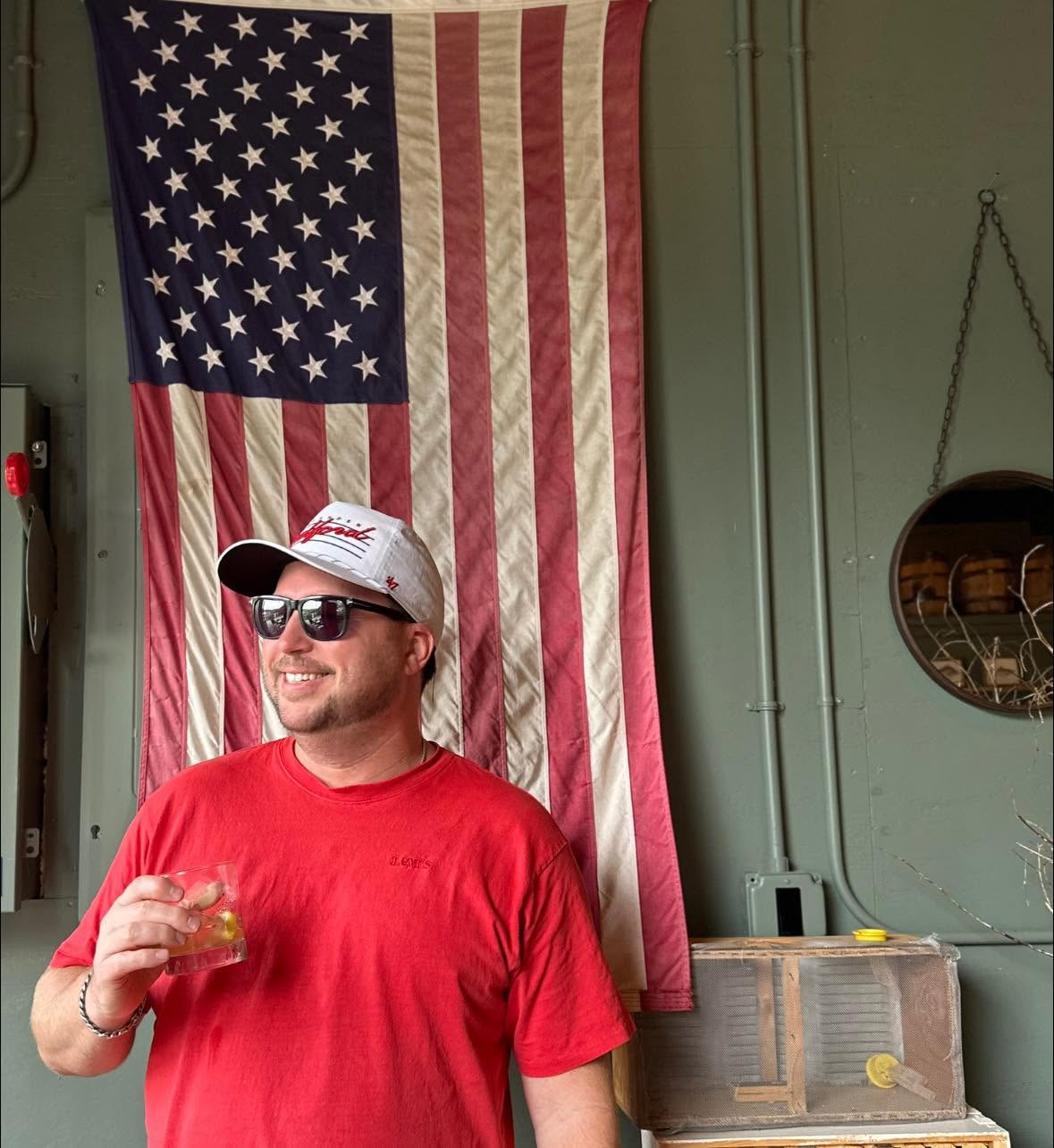 Man wearing sunglasses, a cap, and a red shirt, smiling, holding a drink, standing in front of an American flag.