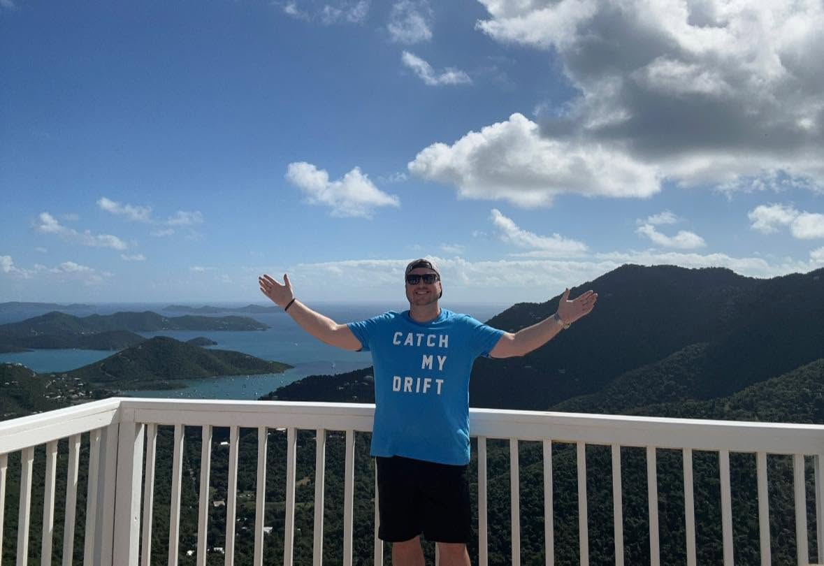 Person standing on a balcony with arms outstretched, scenic mountains and sky in background.