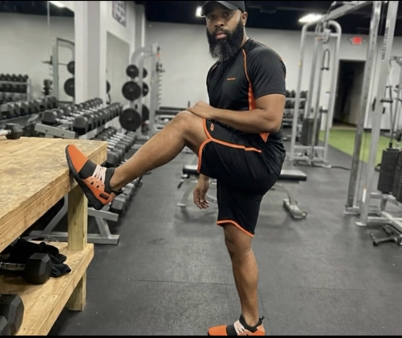 Man in gym stretching leg on wooden bench, surrounded by workout equipment, wearing black and orange sportswear.