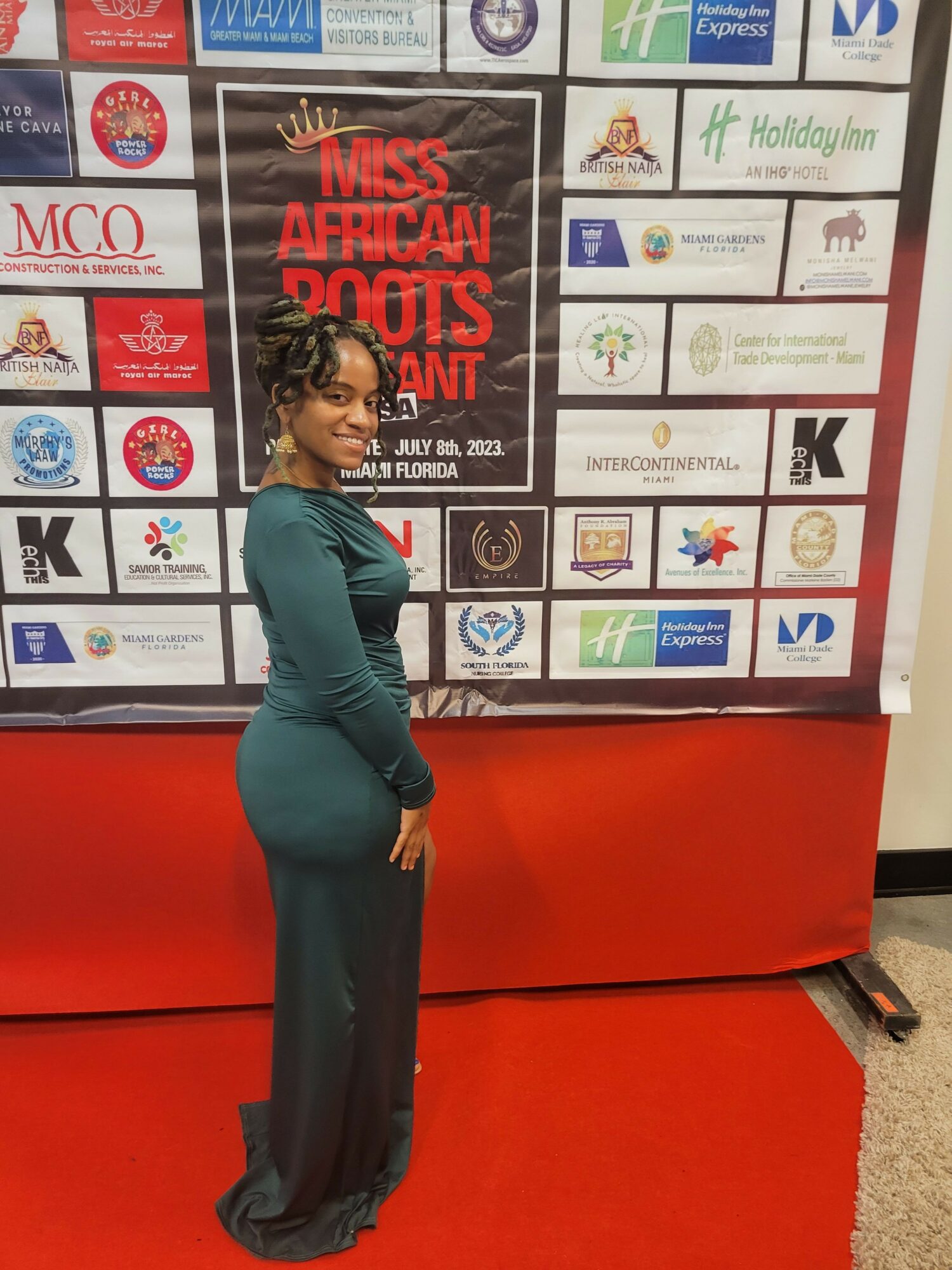 Woman in teal dress standing sideways in front of a display board with various logos and text, red carpet beneath her.