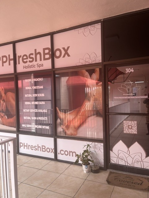 Glass door and window of a spa with a sign, a potted plant, and a doormat outside, reflected in the glass.
