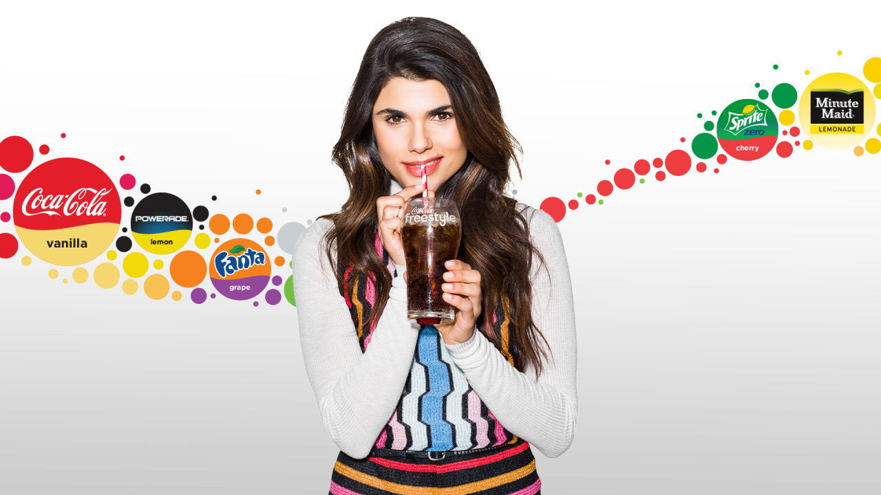Young woman with long dark hair holding a glass of soda, smiling, with colorful brand logos in background.