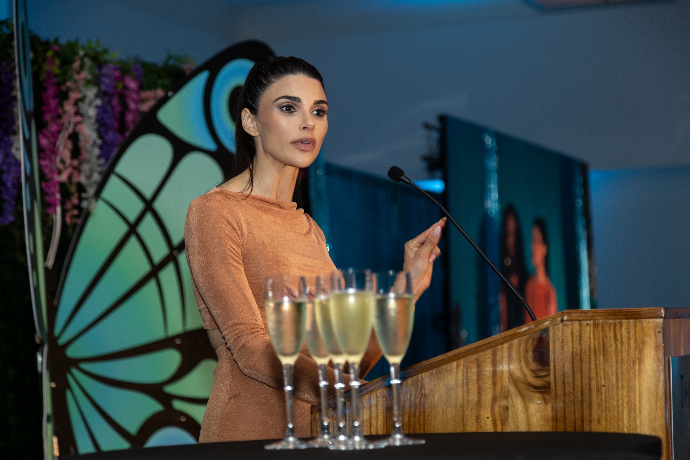 Woman speaking at a podium with a butterfly-shaped backdrop and floral decorations, holding a microphone, in a formal event setting.