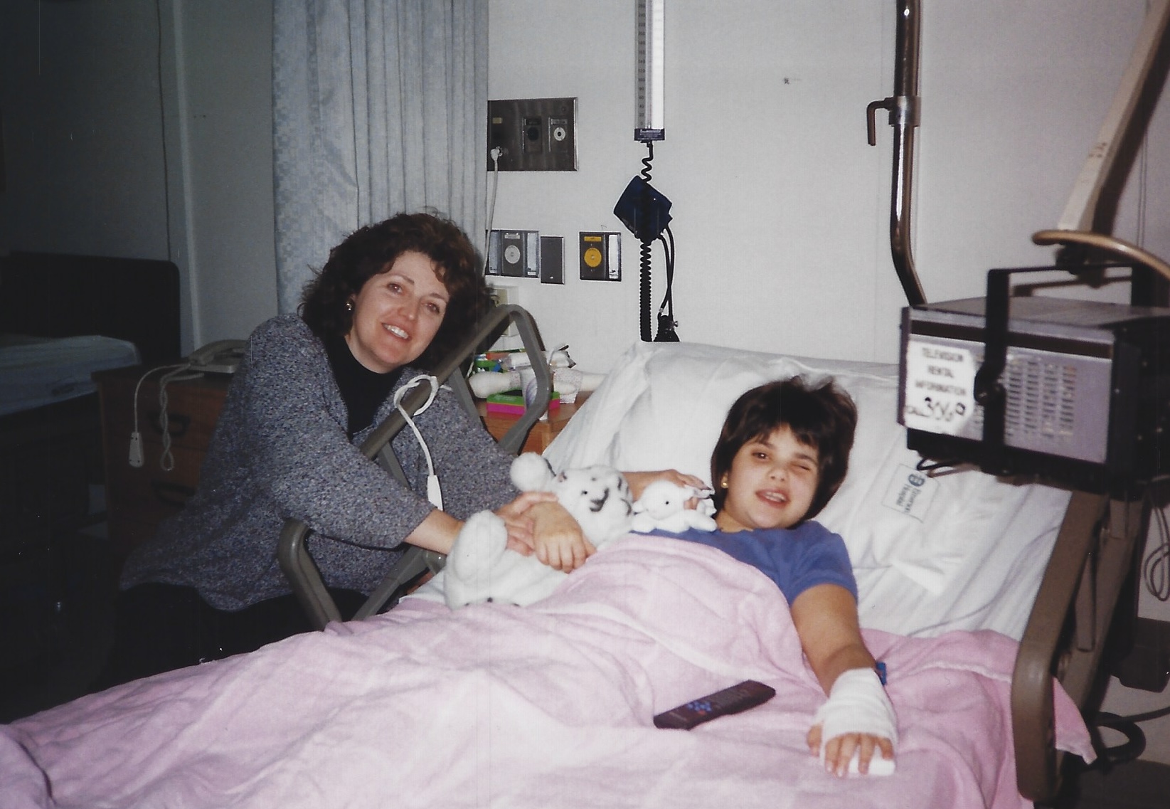 Two women in a hospital room, one lying in bed with a bandaged hand, the other sitting beside her, smiling and holding hands.