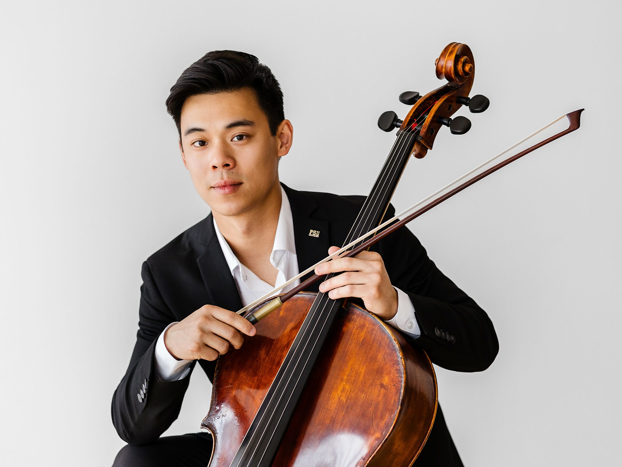 Young man in formal attire holding a cello and bow, looking at the camera, against a plain light background.