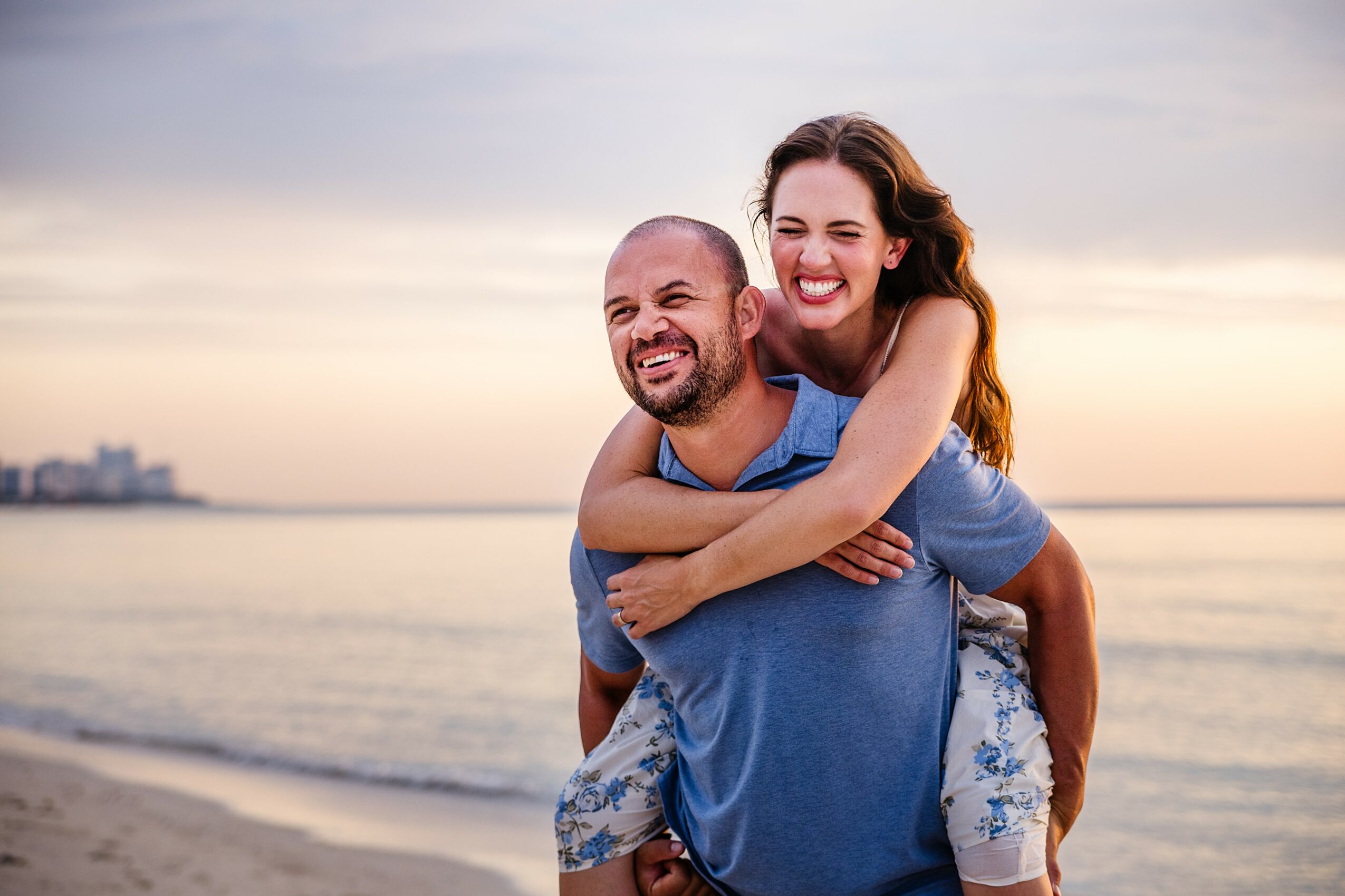 A smiling woman with long hair is on a man's back, both enjoying a beach sunset. The ocean and sky are in the background.