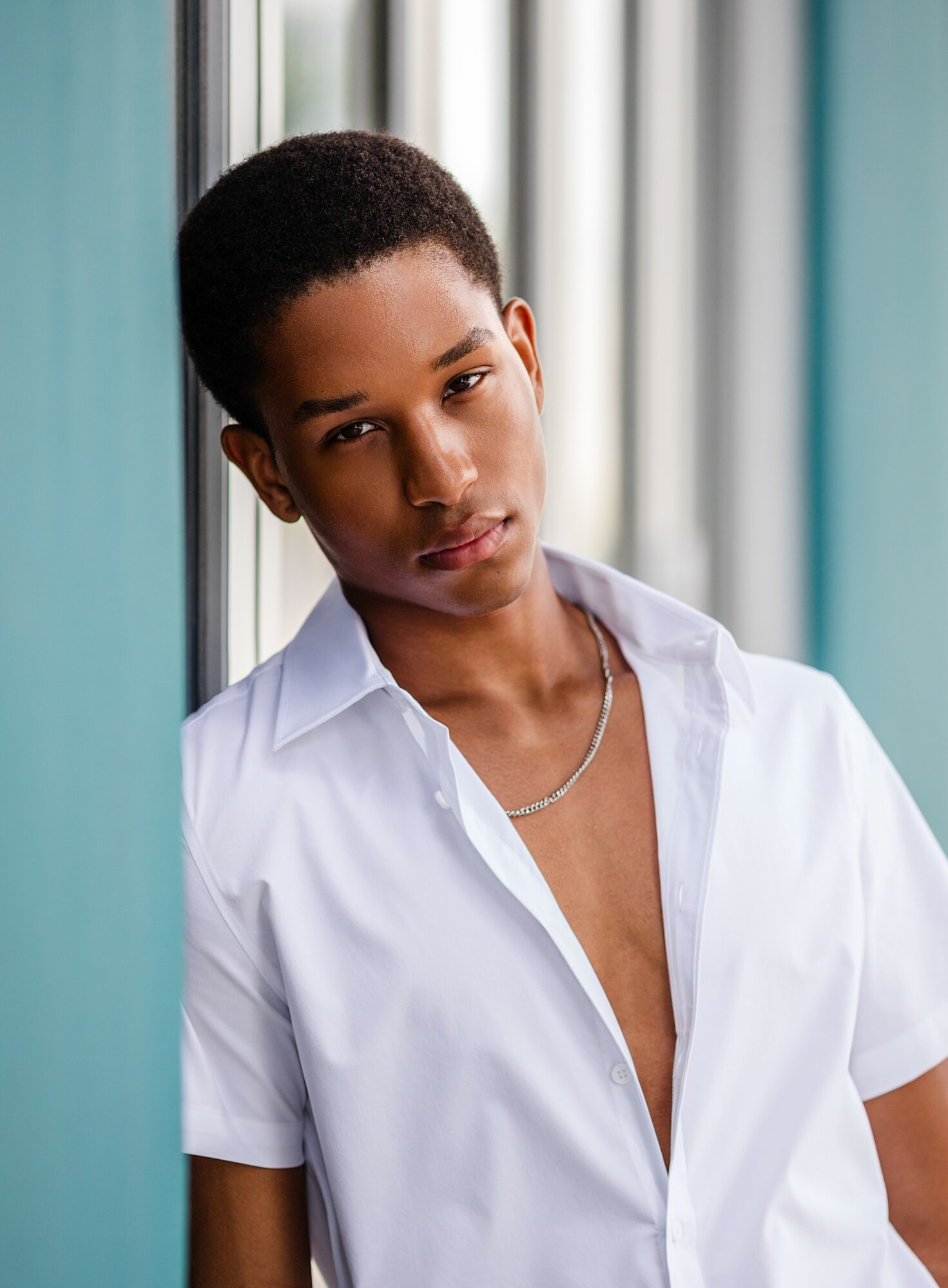 Young person with short hair leaning against a window, wearing a white shirt partially unbuttoned, looking at the camera.