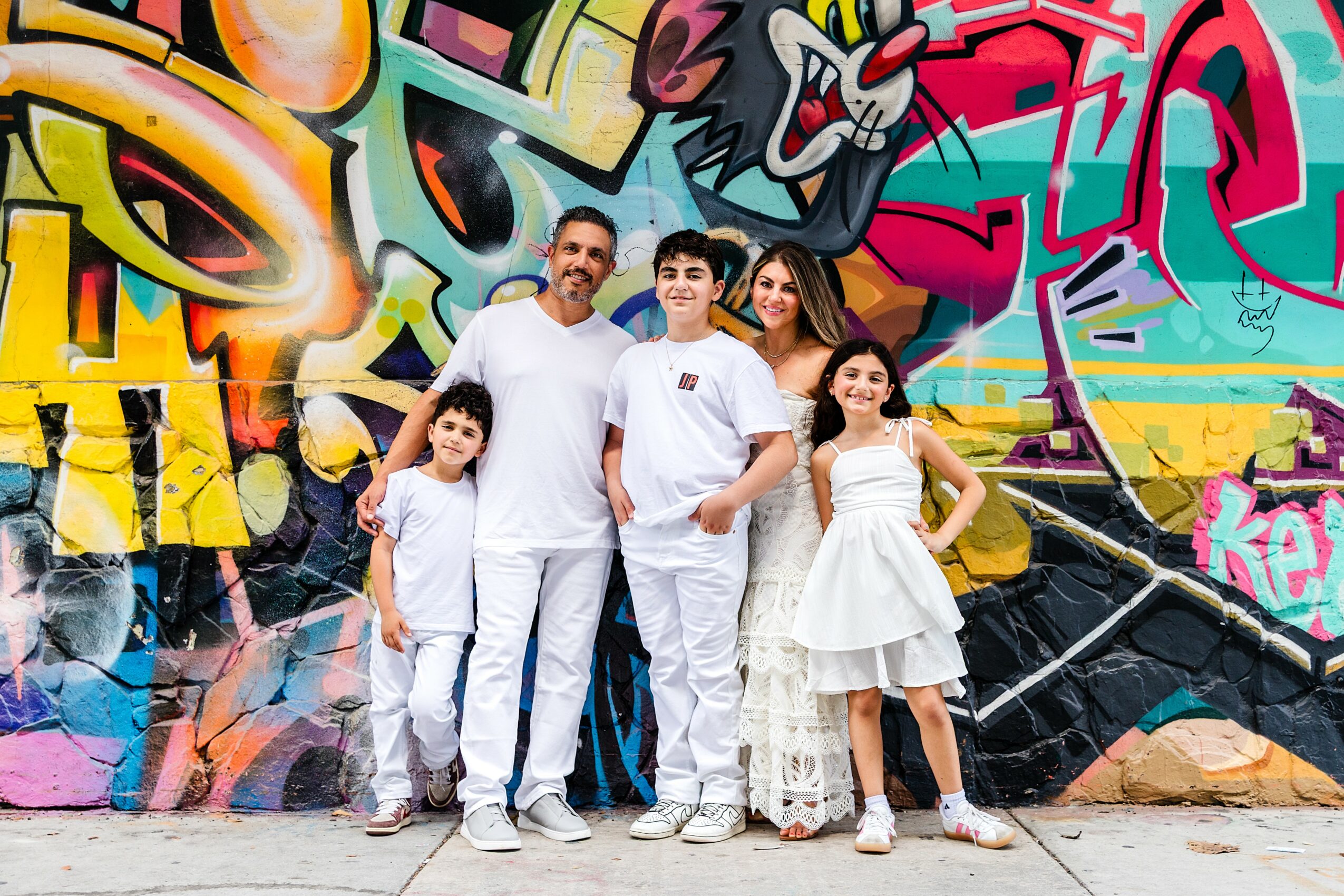 Family of five standing in front of colorful graffiti wall, smiling, with two children and three adults, all dressed in white and casual clothing.
