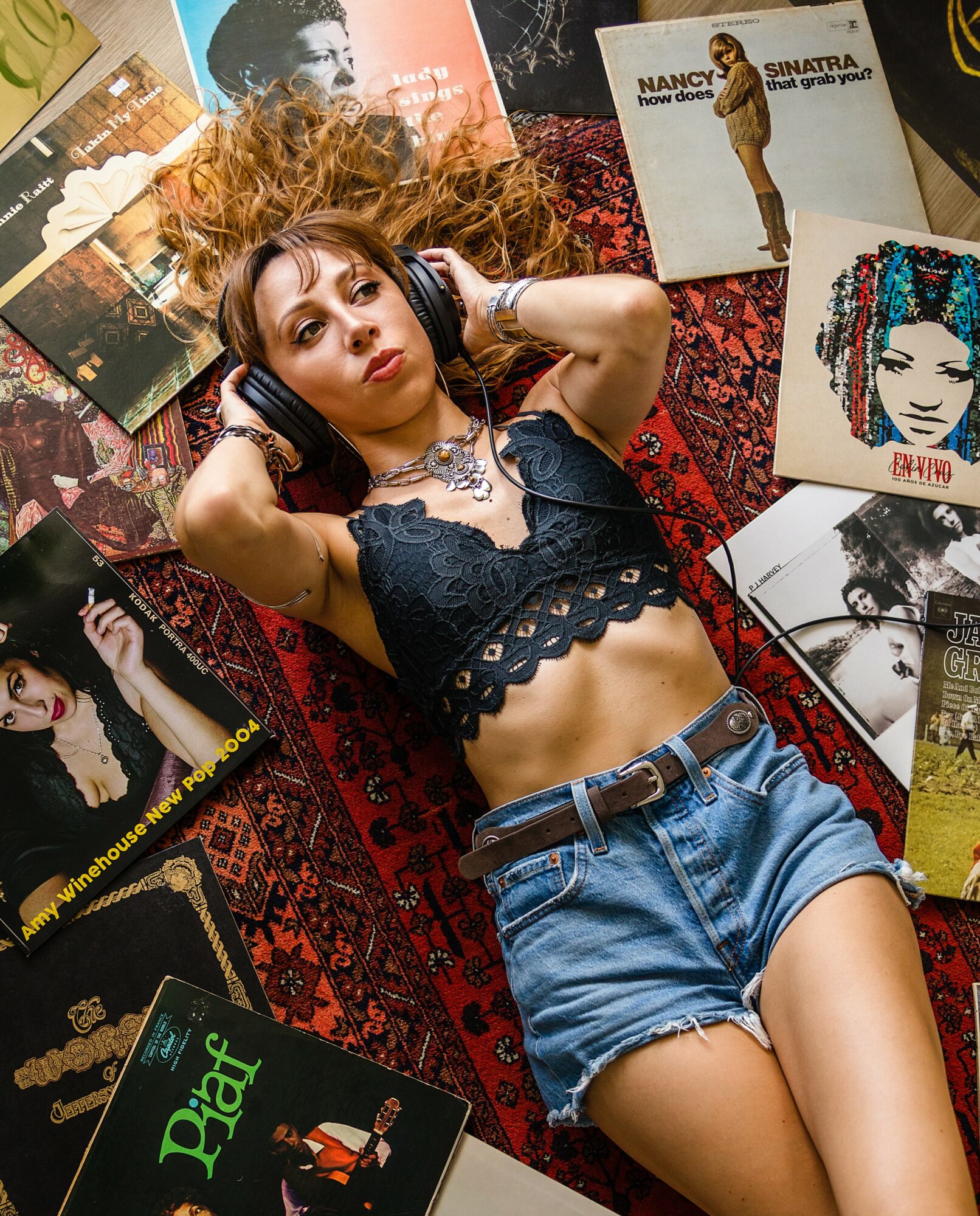 Young woman with curly hair lying on a patterned surface, wearing headphones, a black lace crop top, and denim shorts, surrounded by photographs and artwork.