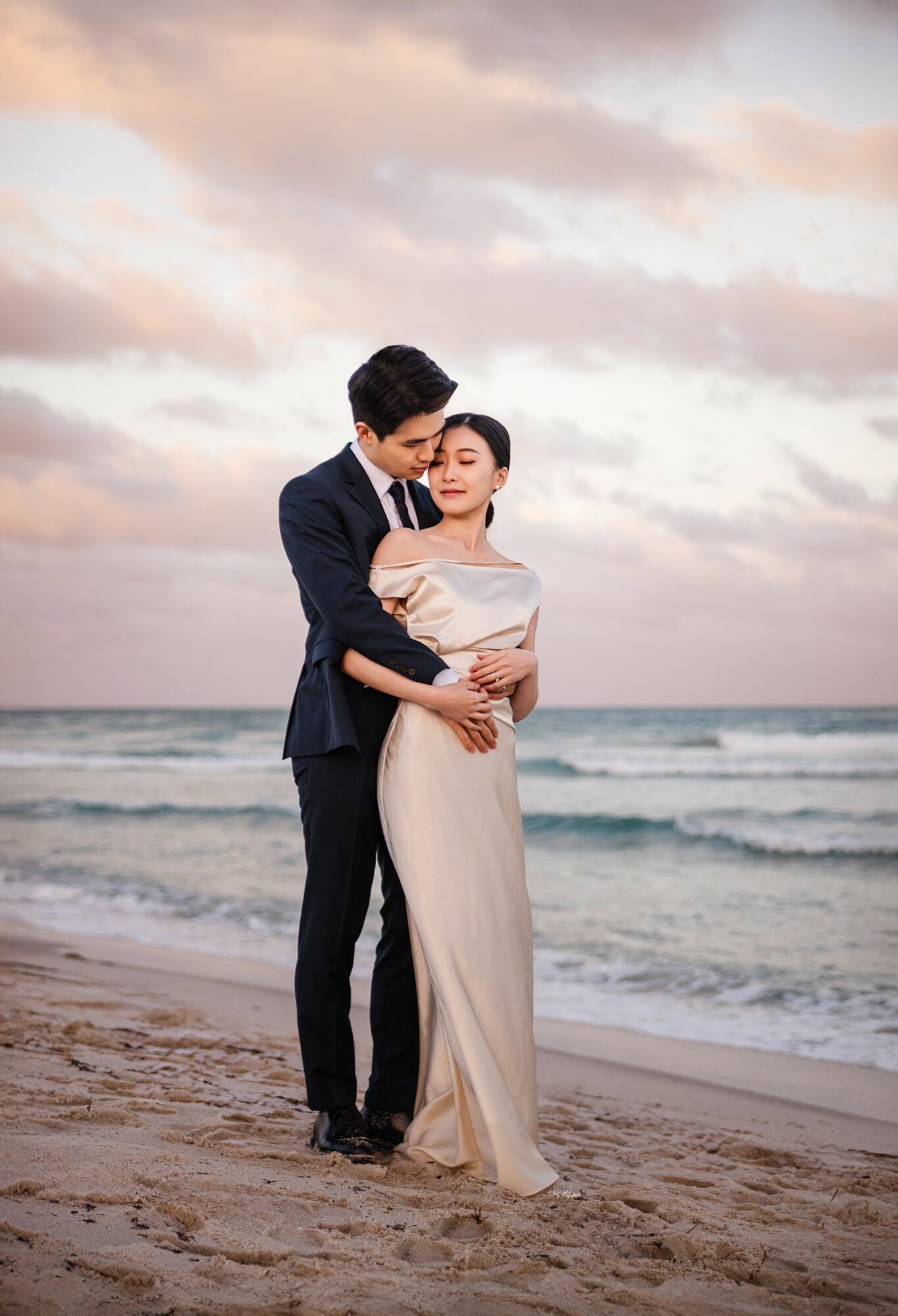 A couple in formal attire embracing on a beach during sunset, with ocean waves and cloudy sky in background.