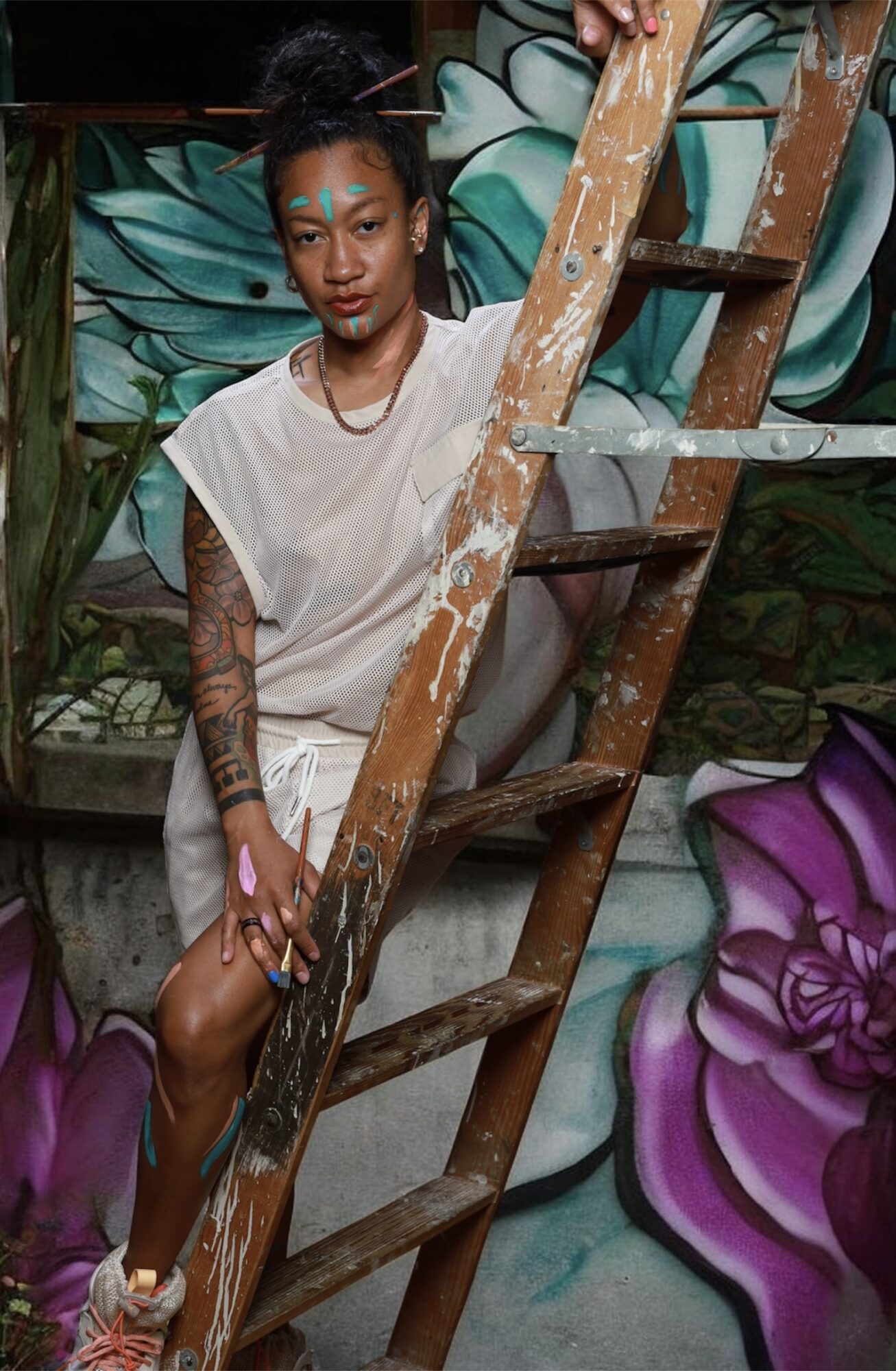 Young woman with tattoos and colorful face paint stands on a ladder in front of a floral mural.