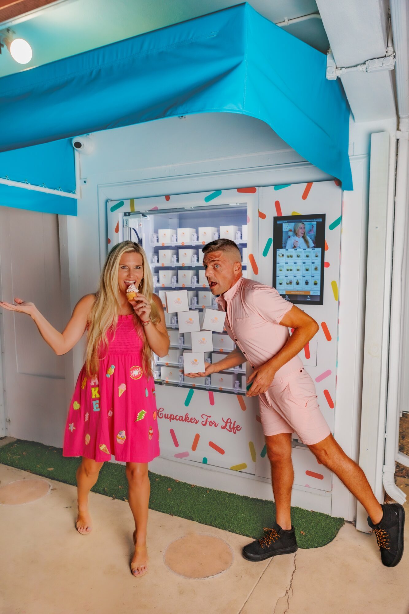 Two people, a woman in a pink dress and a man in pink shorts and black shoes, stand in front of a colorful display at an event.