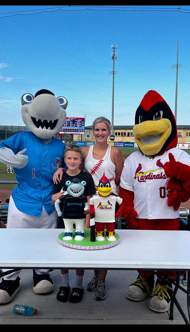 Two children with two mascots, one in a blue costume and the other in a Cardinals uniform, standing outdoors with a table.