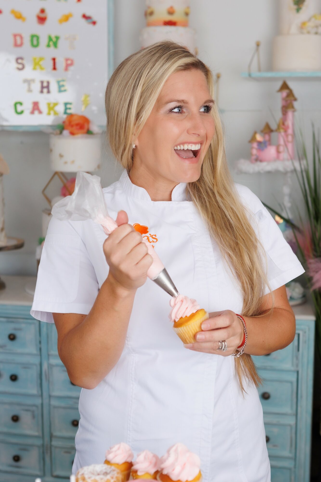 Woman with long blonde hair smiling, holding a cupcake with pink frosting and a piping bag, in a decorated kitchen.