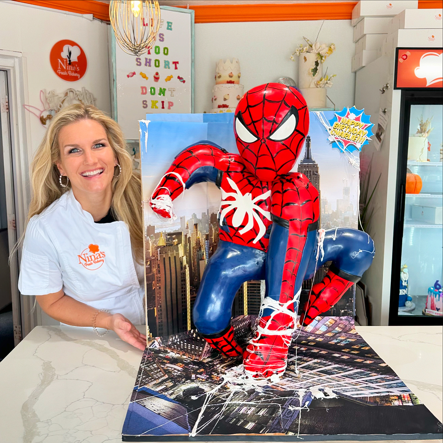 Woman smiling next to a Spider-Man balloon on a cityscape backdrop, on a white table in a decorated room.