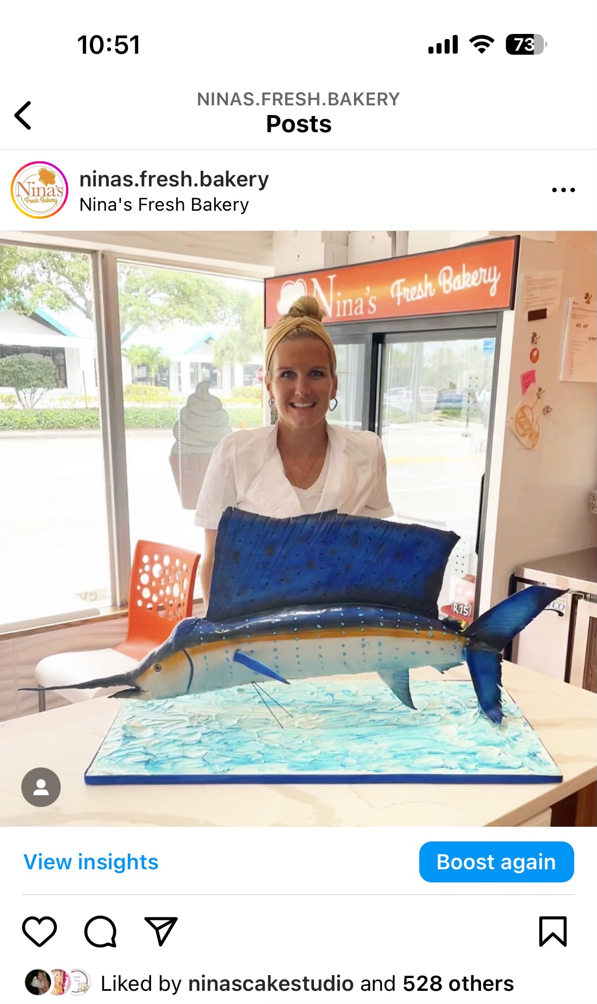 Woman smiling behind a cake shaped like a fish, inside a bakery with large windows and orange signage.