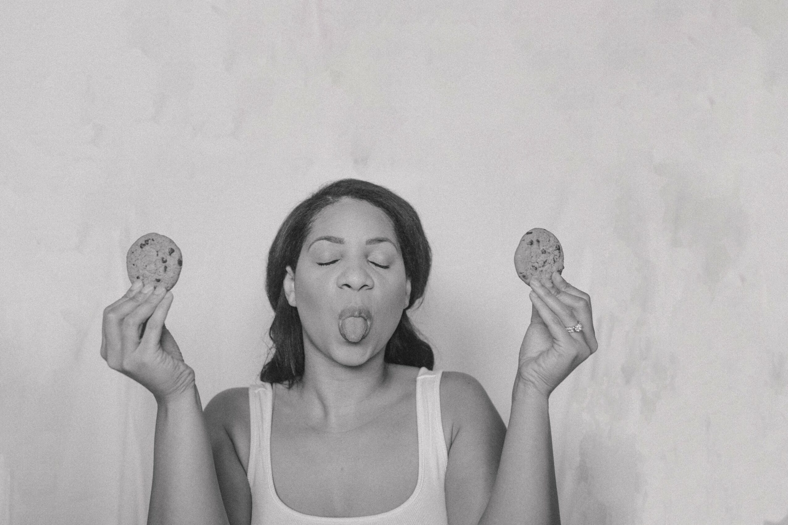Woman with eyes closed and tongue out holding cookies in both hands, against a plain background.