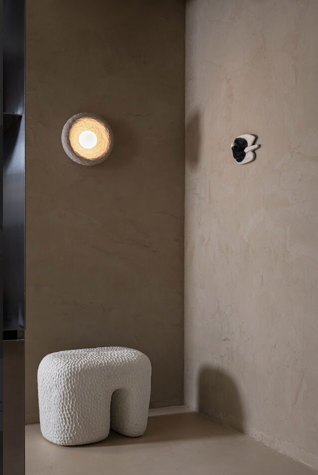 Upright image of a wall with a round light fixture, a small black object, and a textured white stool.