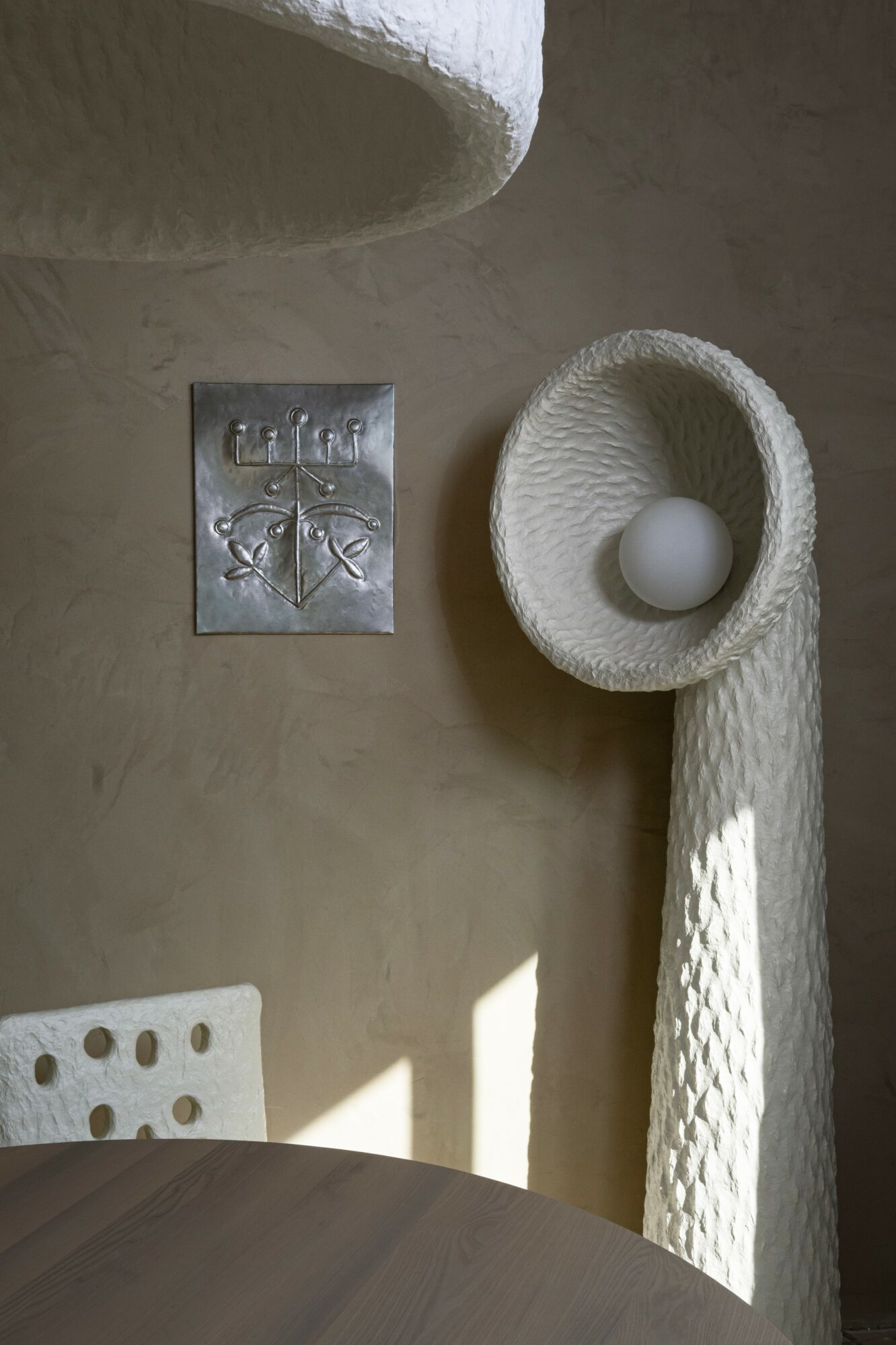 Decorative wall with a metal plaque, a textured white sculpture with a ball, and a shadowed table edge.
