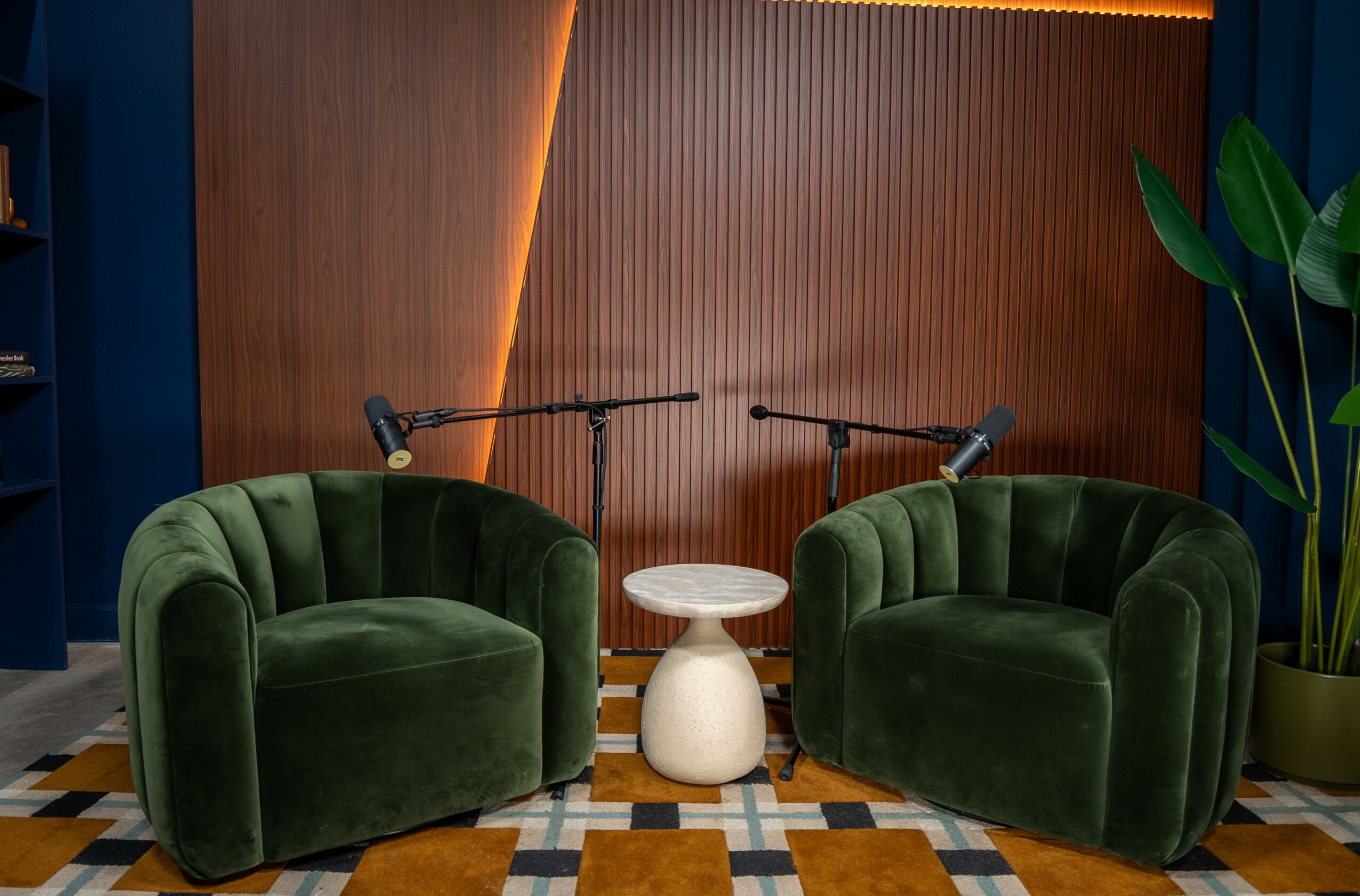 Two green velvet armchairs with a small white side table between them, set against a wooden wall with lighting, and a large plant on the right.