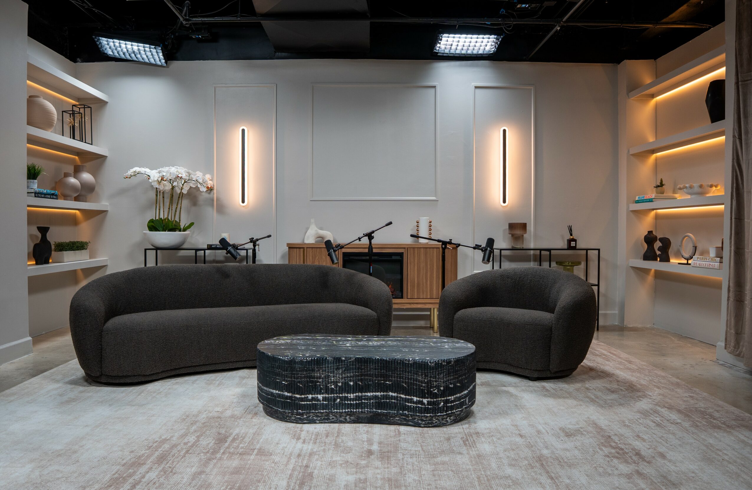 Modern room with two black sofas, a black marble coffee table, and shelves with decorative items, illuminated by wall lights.