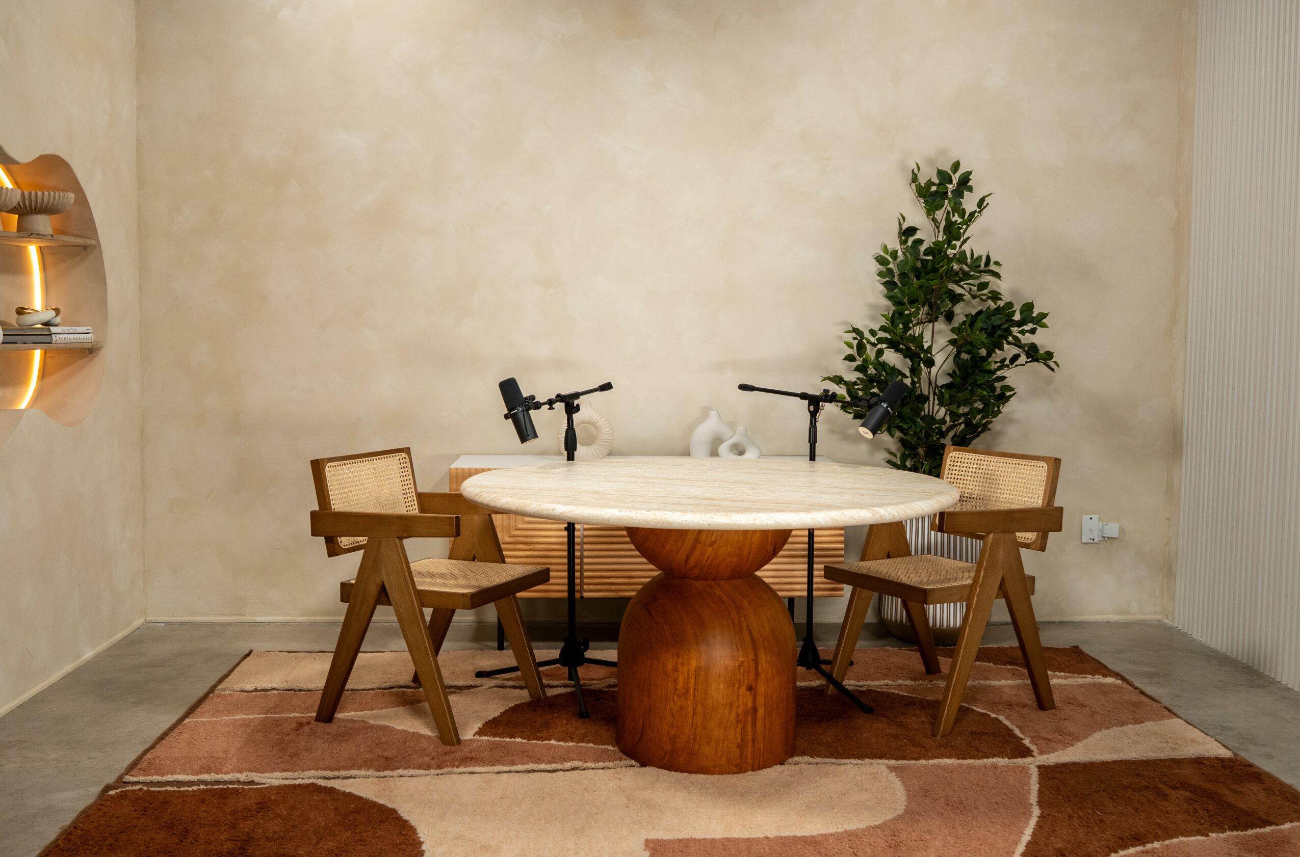 Round table with four chairs, two small microphones, a potted plant, and a rug in a room.