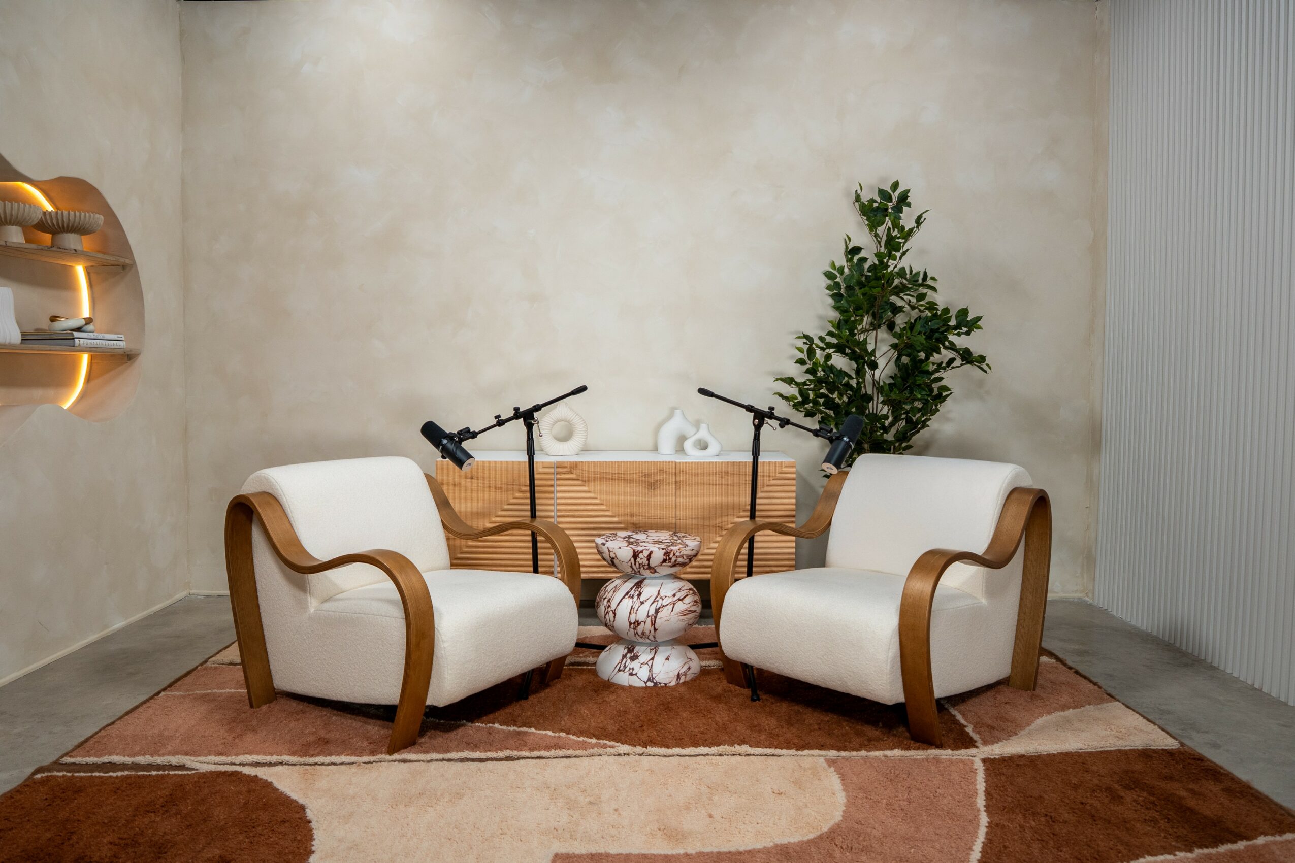 Two white armchairs with wooden accents face each other in a room with a beige wall, a small table, and a plant.
