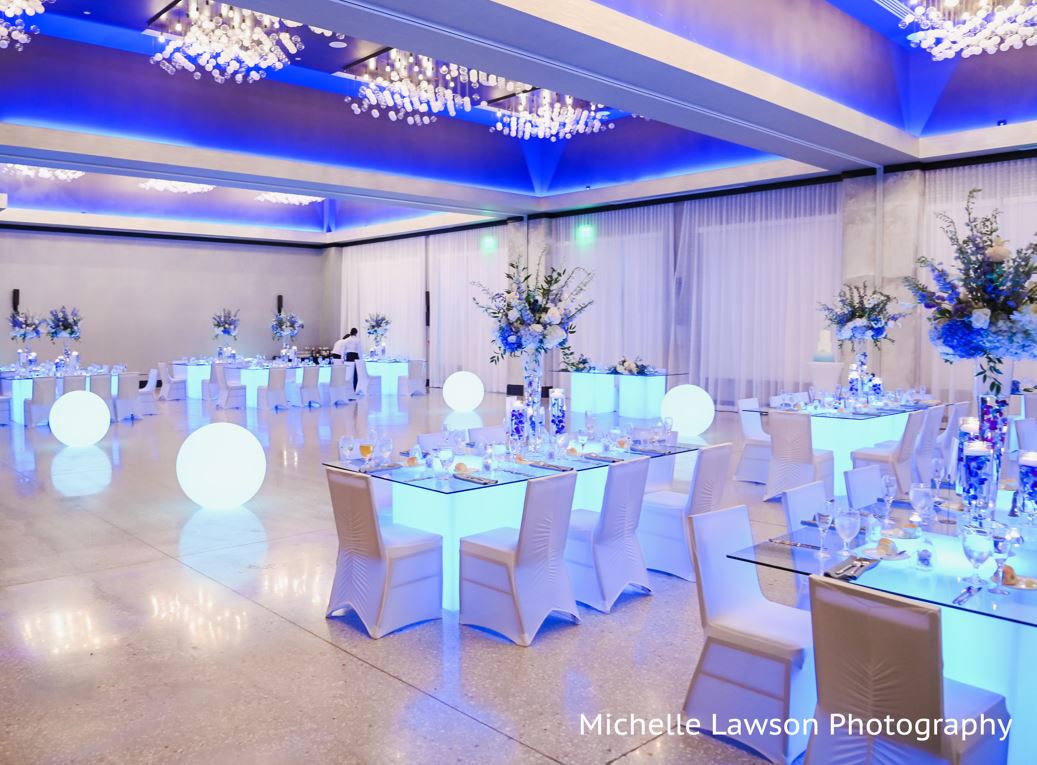 Elegant event space decorated with blue lighting, white chairs, round tables with floral centerpieces, and glowing orbs, ready for a celebration.