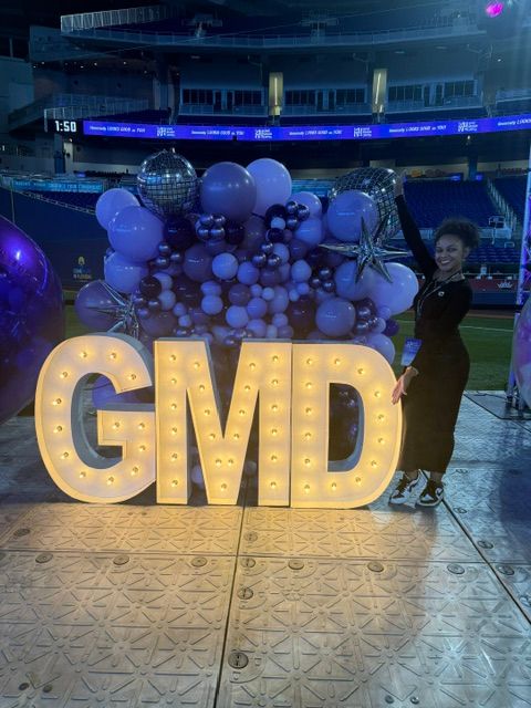 Illuminated sign spelling 'GAY' with balloons and decorations in the background, on a tiled floor.