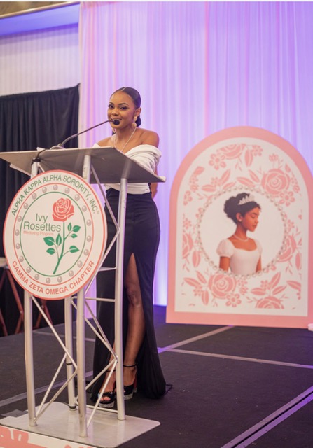 Woman speaking at a podium with a Rosettes logo, standing in front of a pink and purple backdrop and a decorative portrait of a girl.