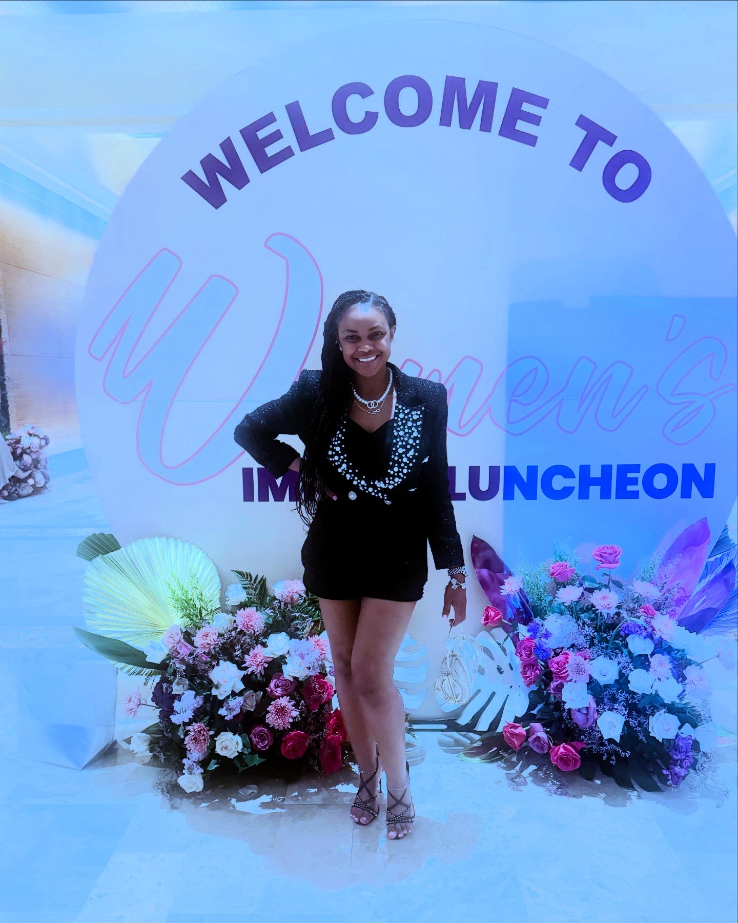 Woman in black dress and pearl necklace standing in front of a floral display and a sign that says 