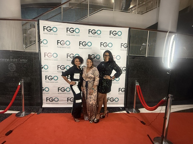 Three women standing on a red carpet in front of a backdrop with FGO logos, posing for a photo.