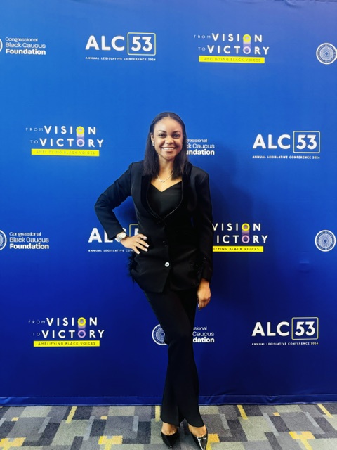 Woman standing in front of a blue backdrop with logos and text, smiling, wearing a black outfit and blazer.