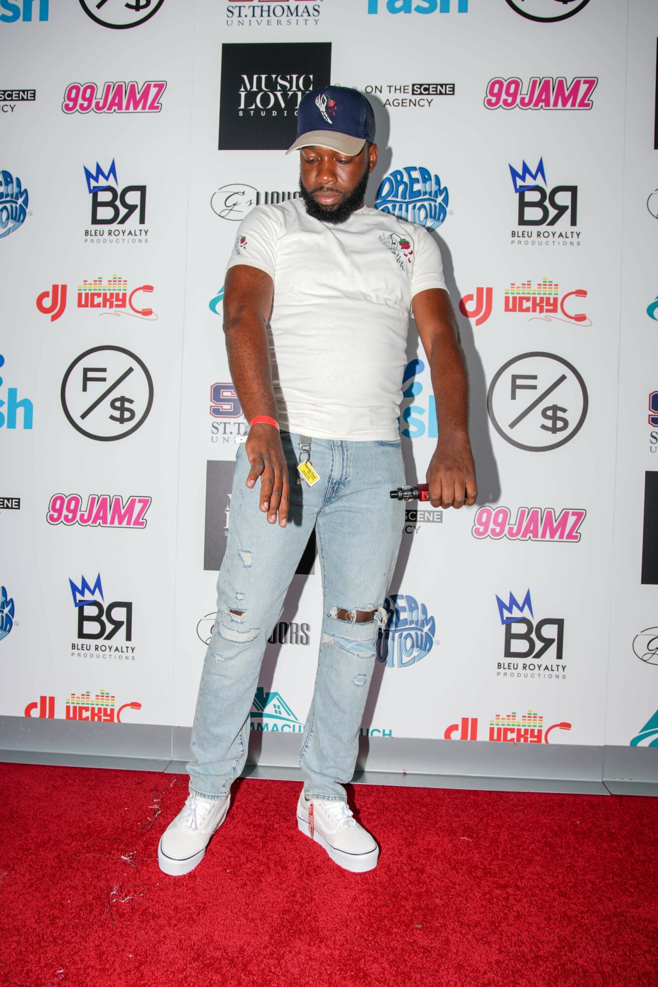 Man in white t-shirt, ripped jeans, white sneakers, and a blue cap standing on red carpet in front of a promotional backdrop.