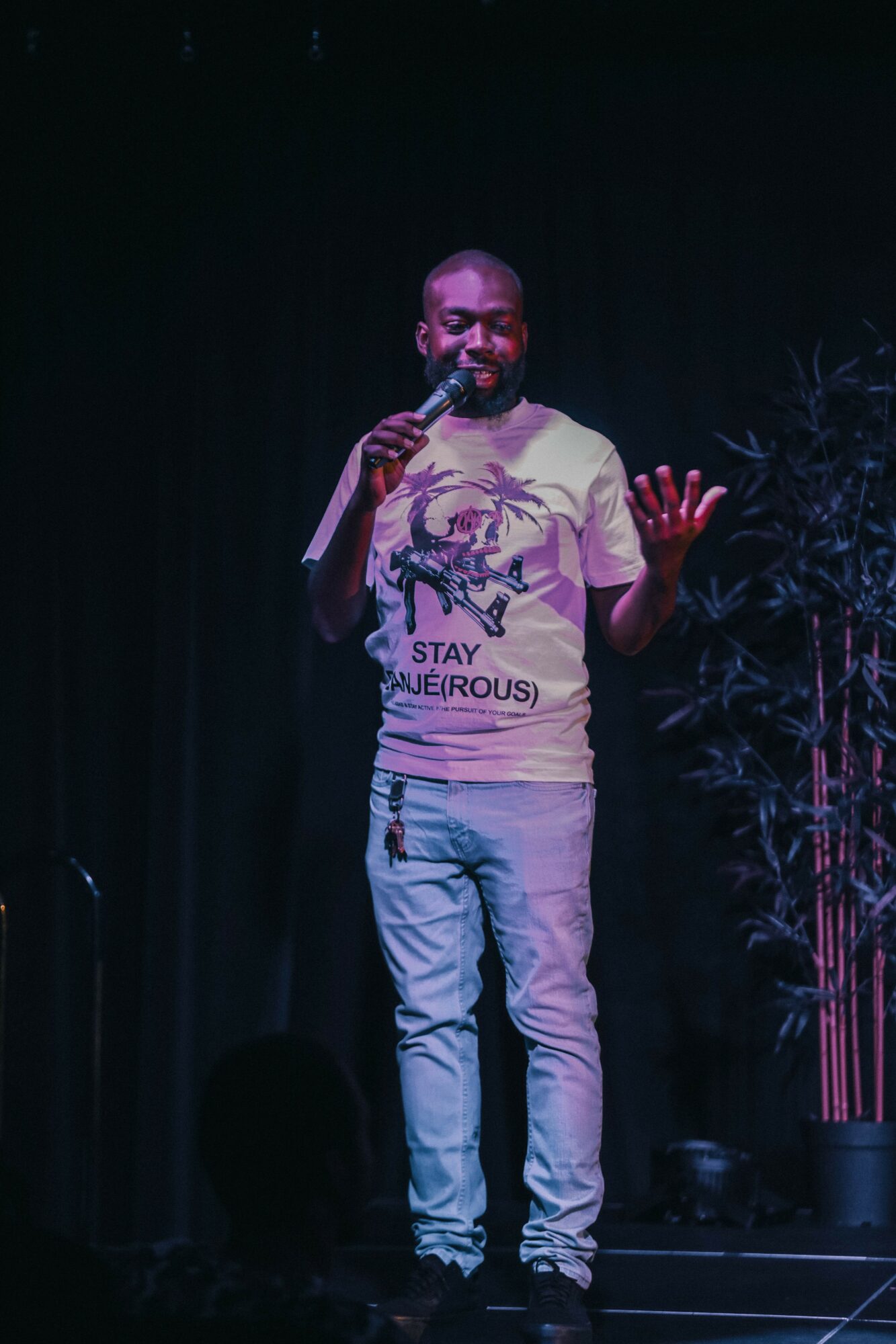 Man with beard holding microphone on stage, wearing a graphic t-shirt and jeans, gesturing with his left hand.