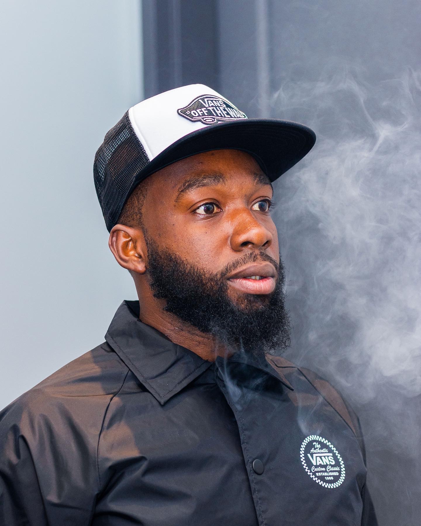 Man with a beard wearing a black shirt and a black and white cap, looking to the side, with smoke nearby.